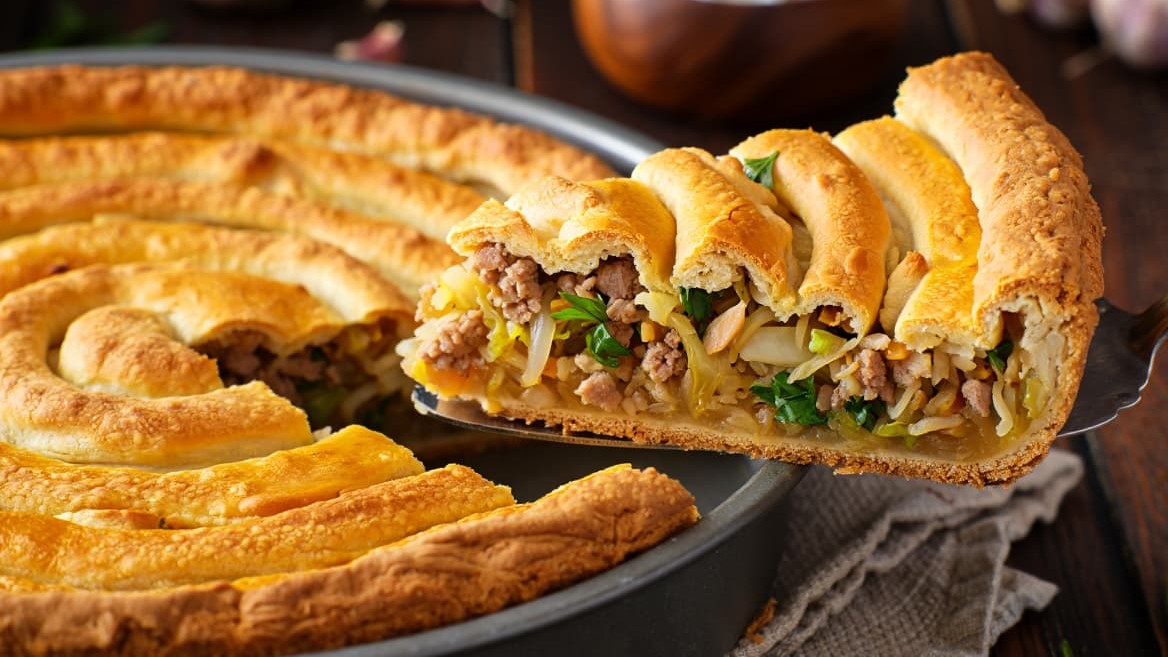 A rustic, golden-brown Serbian savory pie called 'savijača', spiral-shaped in a traditional round metal baking pan. The crust is thin, crispy, and flaky with a slight shine from butter and sour cream wash. A large slice is cut and lifted, revealing a rich, moist filling of sautéed shredded cabbage (zelje), browned minced pork, and visible flecks of fresh green parsley and garlic. Warm steam rises slightly. The background is a dark wooden table with a vintage linen cloth, a wooden bowl of sour cream, and some fresh garlic cloves. Professional food photography, warm lighting, high detail.